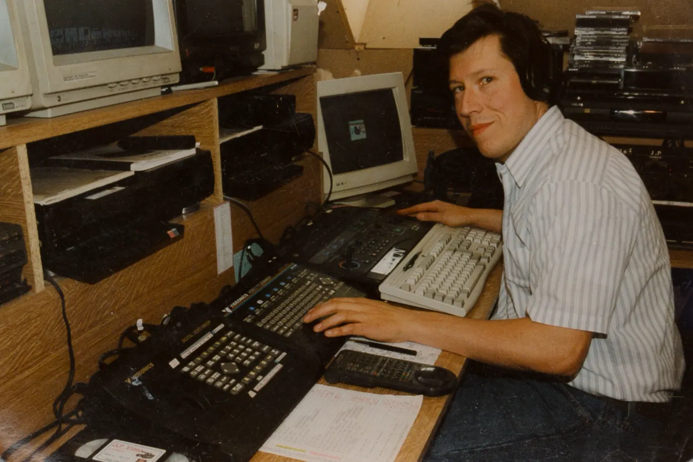 Johan Lenaert met koptelefoon aan zijn montagetafel op zolder, met een rij monitors en een vhs-cassette J.L.P. Video